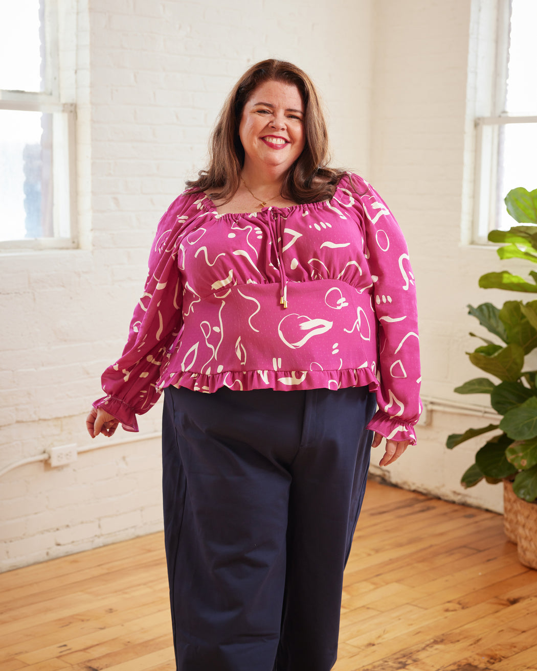 Woman wearing a pink patterned blouse and navy pants in a bright room with a plant. This top is the Briarwood Top sewing pattern from Cashmerette.