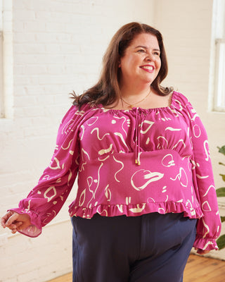Woman wearing a pink blouse with white patterns in a bright room. This top is the Briarwood Top sewing pattern from Cashmerette.