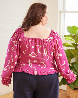 Woman wearing a pink blouse with white patterns in a room with plants. This top is the Briarwood Top sewing pattern from Cashmerette.