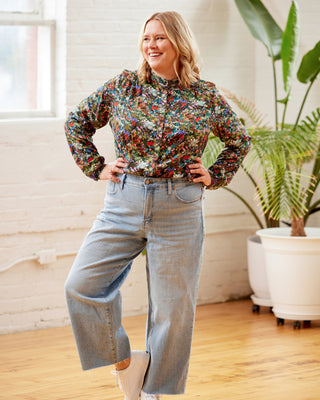 Woman wearing a floral blouse and jeans standing in a room with plants. This top is the Swanton Pussy Bow Blouse sewing pattern from Cashmerette.