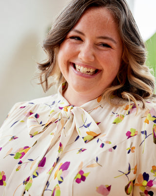 Woman wearing a colorful floral blouse with a blurred background. This top is the Swanton Pussy Bow Blouse sewing pattern from Cashmerette.