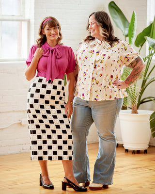 Two women standing indoors, one wearing a pink blouse and black and white checkered skirt, the other in a floral blouse and jeans. This top is the Swanton Pussy Bow Blouse sewing pattern from Cashmerette.