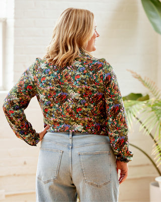 Woman wearing a colorful floral blouse and light blue jeans indoors. This top is the Swanton Pussy Bow Blouse sewing pattern from Cashmerette.