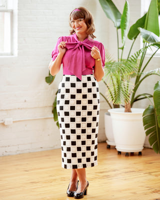 Woman wearing a pink blouse and black and white checkered skirt indoors with plants in the background. This top is the Swanton Pussy Bow Blouse sewing pattern from Cashmerette.