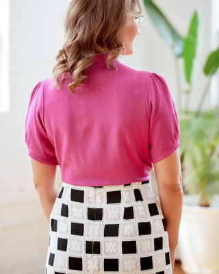 Woman wearing a pink top and black and white checkered skirt indoors. This top is the Swanton Pussy Bow Blouse sewing pattern from Cashmerette.
