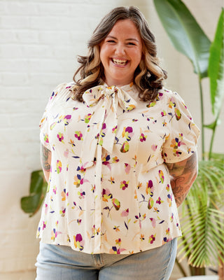 Woman wearing a floral blouse with a white background and plants. This top is the Swanton Pussy Bow Blouse sewing pattern from Cashmerette.