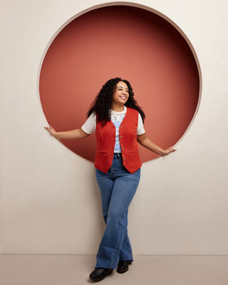 Woman wearing a red vest and blue jeans standing in front of a circular red cutout on a beige wall. This vest is the Hartwell Vest sewing pattern from Cashmerette.