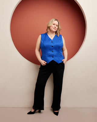 Woman wearing a blue sleeveless top and black pants against a beige wall with a red circular feature. This vest is the Hartwell Vest sewing pattern from Cashmerette.