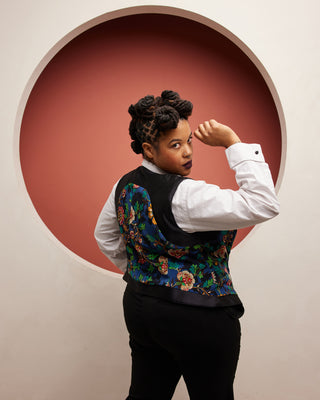 Person wearing a colorful vest with a white shirt and black pants, posing in front of a circular red and beige wall. This vest is the Hartwell Vest sewing pattern from Cashmerette.