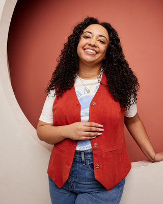 Woman wearing a red vest over a white shirt with a colorful pattern, standing against a warm-toned background. This vest is the Hartwell Vest sewing pattern from Cashmerette.