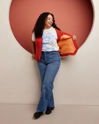 Woman wearing a red jacket, white t-shirt with graphic design, and blue jeans against a beige wall with a large circular cutout. This vest is the Hartwell Vest sewing pattern from Cashmerette.