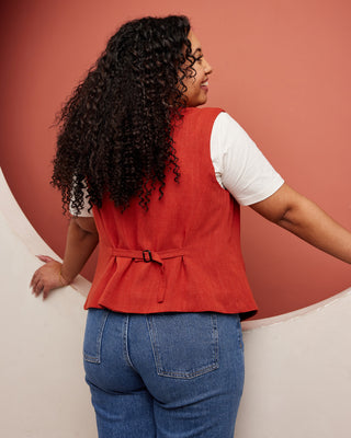 Person wearing a red vest over a white shirt and blue jeans against a pink background. This vest is the Hartwell Vest sewing pattern from Cashmerette.