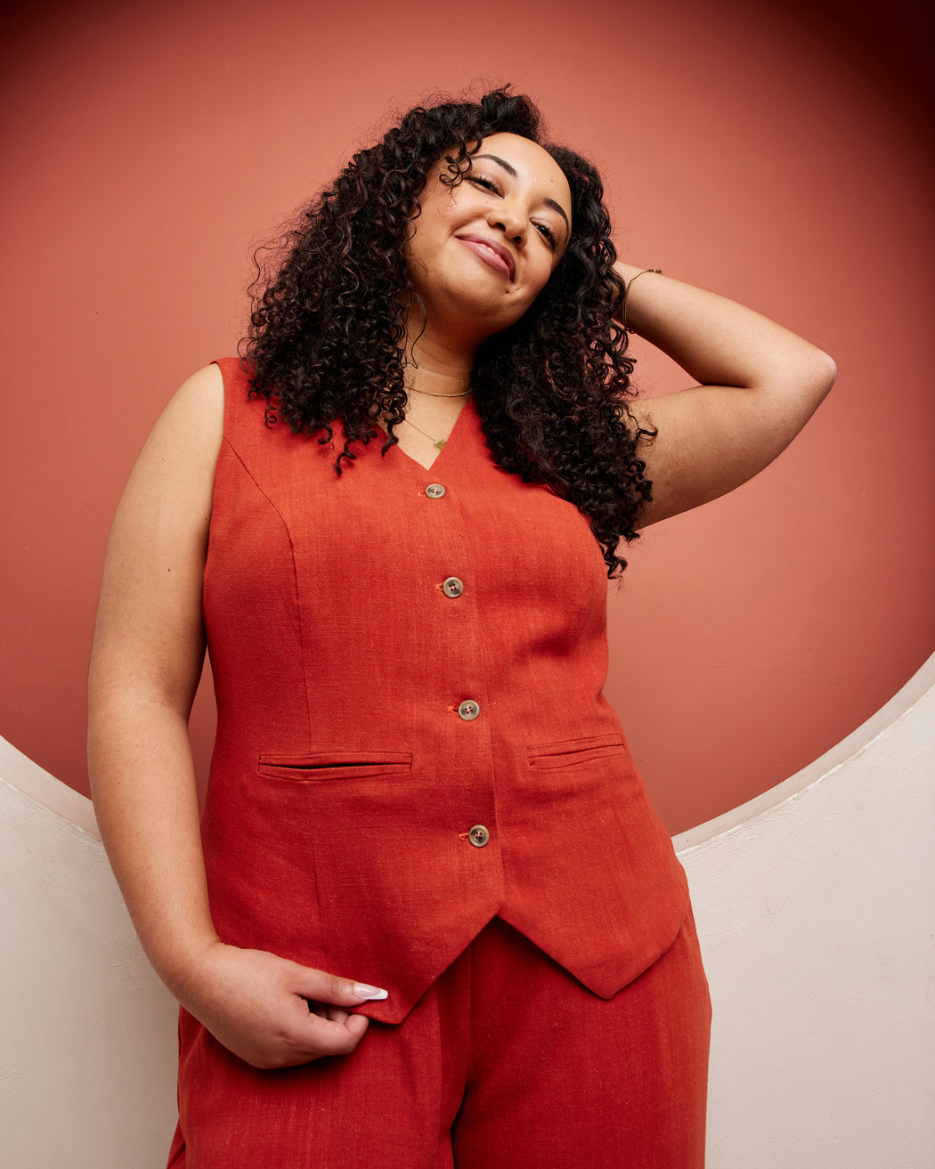 Woman wearing a red sleeveless top and pants against a pink background. This vest is the Hartwell Vest sewing pattern from Cashmerette.