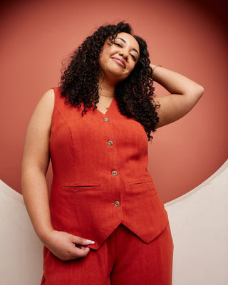 Woman wearing a red sleeveless top and pants against a pink background. This vest is the Hartwell Vest sewing pattern from Cashmerette.