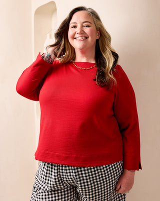 A woman wearing a bright red long-sleeve top and black-and-white gingham pants, standing against a warm beige wall. This is the Wellfleet Breton T-Shirt sewing pattern from Cashmerette.