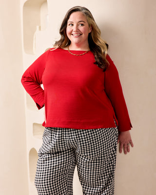 A woman modeling a bright red long-sleeve knit top paired with black and white gingham pants, standing against a beige wall with recessed architectural details. This is the Wellfleet Breton T-Shirt sewing pattern from Cashmerette.