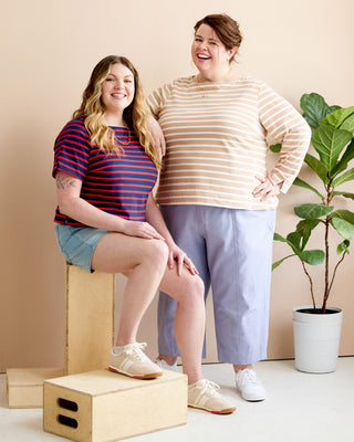 Two women modeling striped long-sleeve tops in a studio setting. One woman sits on a wooden box wearing a navy and red striped shirt with denim shorts, and the other stands wearing a beige and white striped shirt with light blue cropped pants. This is the Wellfleet Breton T-Shirt sewing pattern from Cashmerette.
