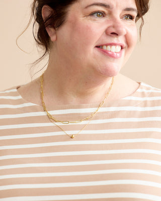 A smiling woman wearing a layered gold necklace over a beige and white striped top, set against a solid light beige background. This is the Wellfleet Breton T-Shirt sewing pattern from Cashmerette.