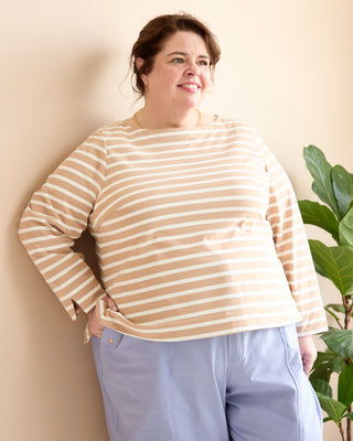 A woman wearing a beige and white horizontally striped long-sleeve top and light blue pants, standing against a beige wall next to a green plant. This is the Wellfleet Breton T-Shirt sewing pattern from Cashmerette.