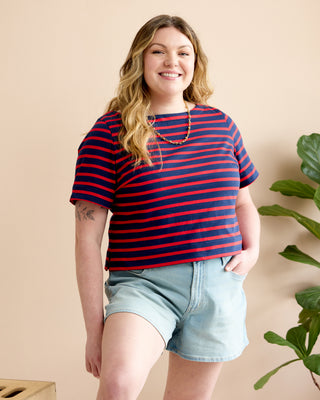 A woman modeling a short-sleeve striped top with navy and red horizontal stripes, paired with light blue denim shorts, against a warm beige background with a green plant on the side. This is the Wellfleet Breton T-Shirt sewing pattern from Cashmerette.