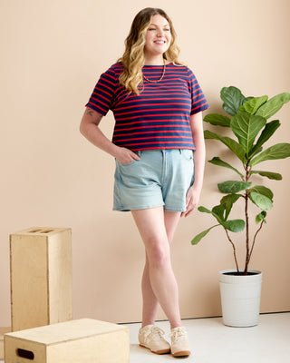 A woman stands in a studio wearing a navy and red horizontally striped short-sleeve t-shirt, light blue denim shorts, and beige sneakers. She is posed next to a potted fiddle-leaf fig plant and wooden crates against a solid beige background. This is the Wellfleet Breton T-Shirt sewing pattern from Cashmerette.