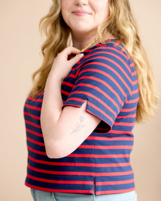 A woman wearing a short-sleeve striped top with navy blue and red horizontal stripes, paired with light blue jeans, set against a solid beige background. This is the Wellfleet Breton T-Shirt sewing pattern from Cashmerette.