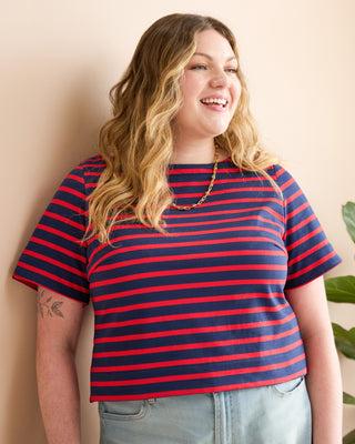 A woman with wavy blonde hair wearing a red and navy blue horizontally striped short-sleeve crop top, paired with light blue jeans, posing against a beige wall. This is the Wellfleet Breton T-Shirt sewing pattern from Cashmerette.