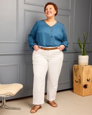 Woman wearing a blue blouse and white pants standing in a room with gray walls and a wooden plant stand. This is the Inman Top & Dress sewing pattern from Cashmerette.
