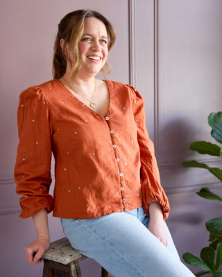 Woman wearing an orange blouse with pearl embellishments sitting on a stool against a pink wall. This is the Inman Top & Dress sewing pattern from Cashmerette.