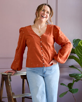 Woman wearing an orange blouse and light blue jeans standing on a wooden stool against a pink wall. This is the Inman Top & Dress sewing pattern from Cashmerette.