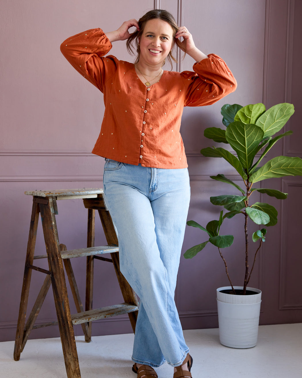 Woman in an orange blouse and light blue jeans standing next to a plant and wooden stool against a pink wall. This is the Inman Top & Dress sewing pattern from Cashmerette.