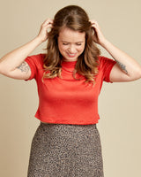 Woman wearing a red shirt and patterned skirt adjusting her hair against a beige background. This t-shirt is the Concord T-Shirt sewing pattern from Cashmerette.