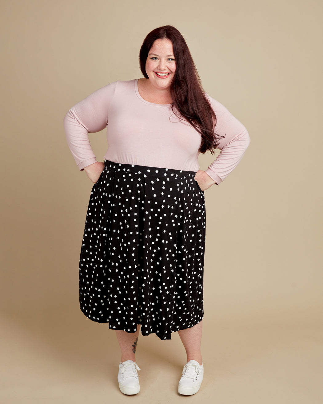 Woman wearing a pink top and black polka dot skirt on a beige background. This t-shirt is the Concord T-Shirt sewing pattern from Cashmerette.