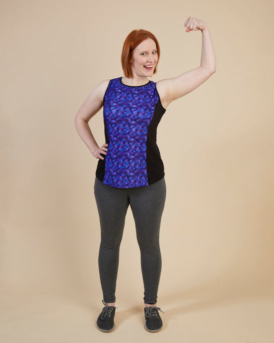 Woman wearing a sleeveless top with a blue pattern and gray leggings, posing against a beige background. This top is the Spruce Workout Tank sewing pattern from Cashmerette.