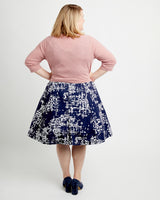 Woman wearing a pink sweater and navy skirt with white pattern on a plain background. This cardigan is the Fuller Cardigan sewing pattern from Cashmerette.