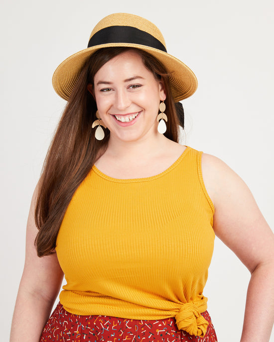 Woman wearing a yellow sleeveless top and a straw hat with a black band on a light gray background. This tank top is the Saybrook Tank sewing pattern from Cashmerette.