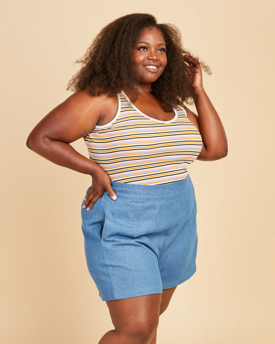Woman wearing a striped tank top and blue shorts against a beige background. This tank top is the Saybrook Tank sewing pattern from Cashmerette.