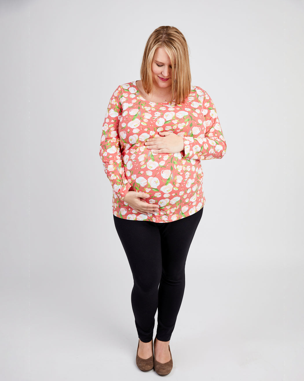 Woman wearing a pink floral top and black pants on a white background. This t-shirt is the Brookline Maternity T-Shirt sewing pattern from Cashmerette.