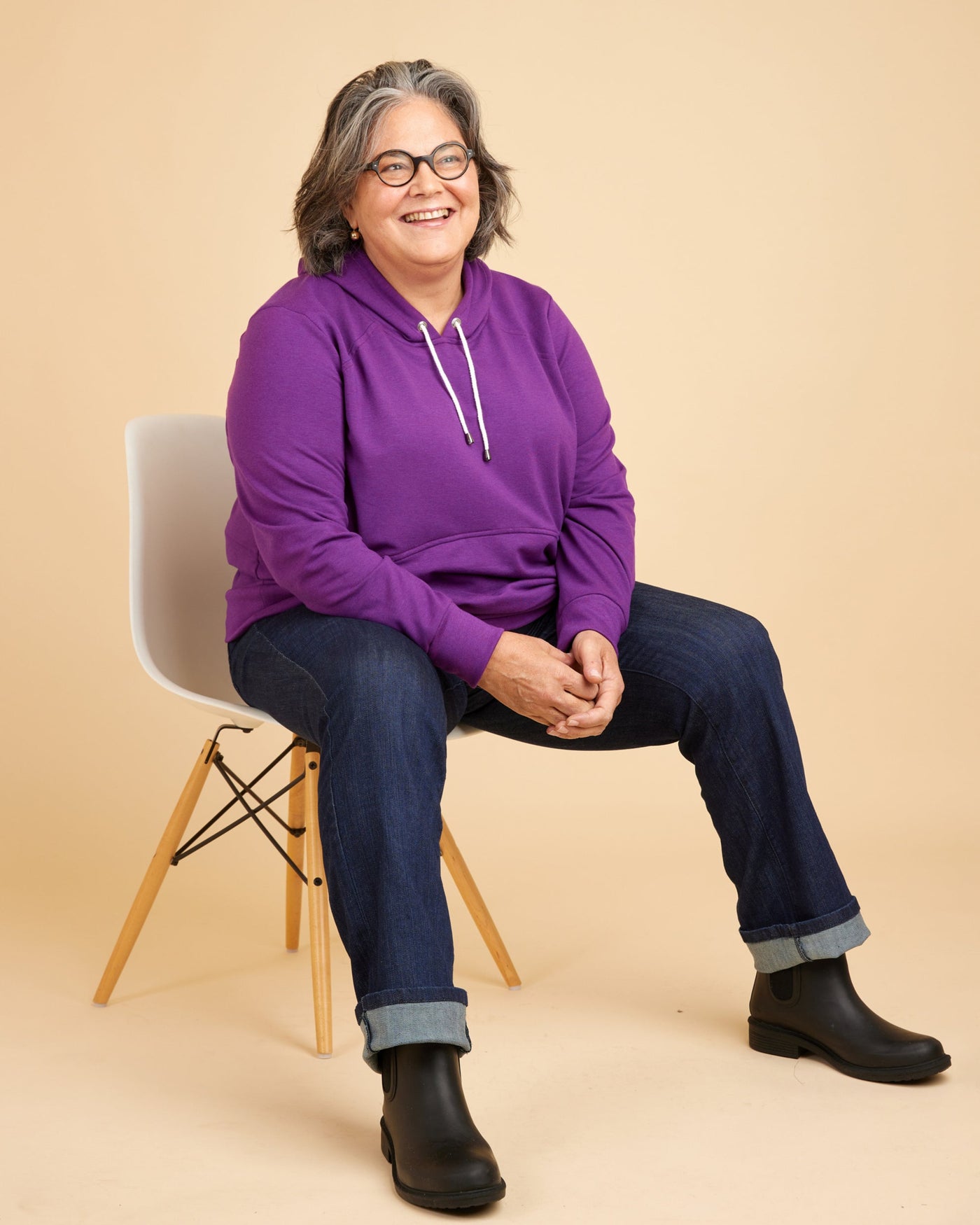 Person wearing a purple hoodie sitting on a chair against a beige background. This hoodie is the Stanton Hoodie sewing pattern from Cashmerette.