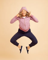 Person wearing a pink jacket and navy pants jumping on a beige background. This hoodie is the Stanton Hoodie sewing pattern from Cashmerette.