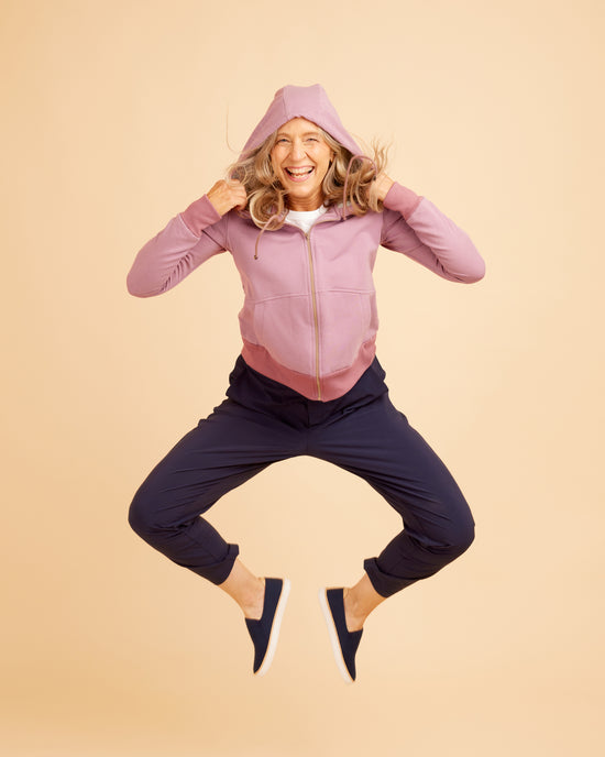 Person wearing a pink jacket and navy pants jumping on a beige background. This hoodie is the Stanton Hoodie sewing pattern from Cashmerette.