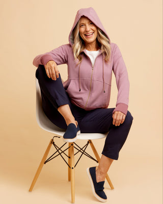 Woman wearing a pink hoodie and navy pants sitting on a white chair against a beige background. This hoodie is the Stanton Hoodie sewing pattern from Cashmerette.