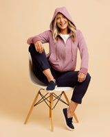 Woman sitting on a chair wearing a pink hoodie and navy pants against a beige background. This hoodie is the Stanton Hoodie sewing pattern from Cashmerette.