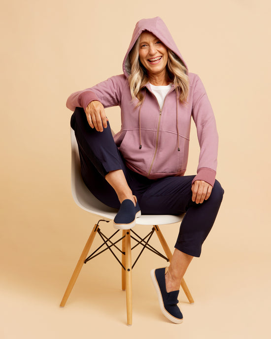 Woman sitting on a chair wearing a pink hoodie and navy pants against a beige background. This hoodie is the Stanton Hoodie sewing pattern from Cashmerette.