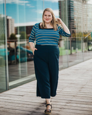 Woman in a striped blue top and dark pants standing on a wooden deck with a cityscape background. This top is the Carlyle square neck t-shirt sewing pattern from Cashmerette.