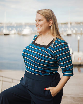 Woman wearing a blue striped shirt and dark pants by a waterfront. This top is the Carlyle square neck t-shirt sewing pattern from Cashmerette.
