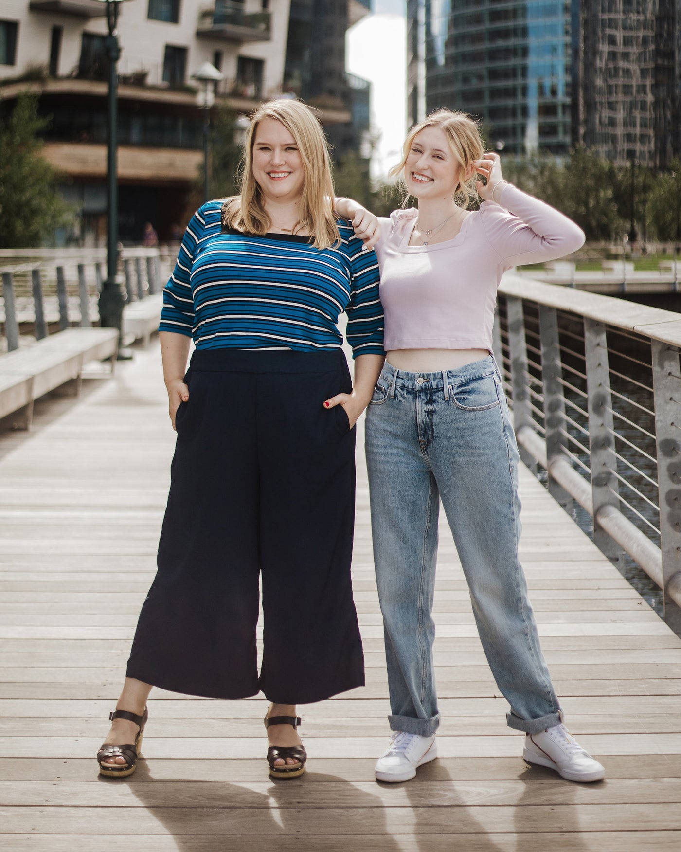 Two women standing on a wooden boardwalk with cityscape in the background. This top is the Carlyle square neck t-shirt sewing pattern from Cashmerette.