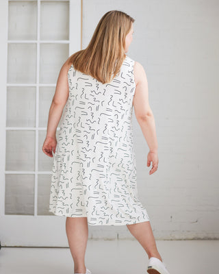 Woman wearing a white dress with black patterns in a minimal indoor setting. This is the Wexford Top & Dress sewing pattern from Cashmerette.