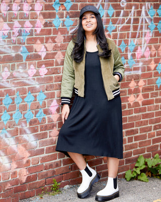Woman wearing a black dress, green jacket, and white shoes against a colorful brick wall. This is the Wexford Top & Dress sewing pattern from Cashmerette.