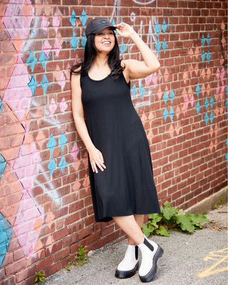 Woman in a black dress and cap standing against a colorful brick wall. This is the Wexford Top & Dress sewing pattern from Cashmerette.